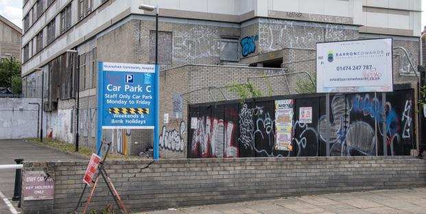 Graffiti-covered wall of Gravesham Community Hospital with signage for staff parking and a nearby business.