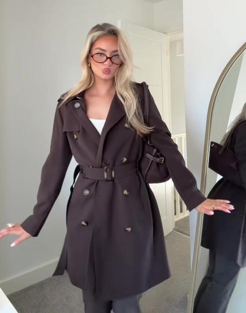 A blonde woman modeling a chocolate brown H&M trench coat with a belt, white top, and holding a brown purse.