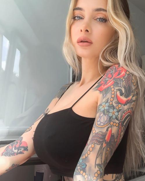 A blonde woman with blue eyes, light makeup, and full arm tattoos wearing a black top.