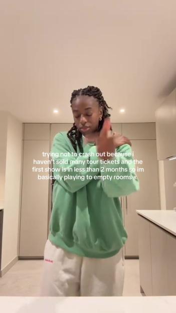 A woman in a green sweatshirt and white sweatpants gestures in a kitchen, with text overlay about her struggles to sell tour tickets.