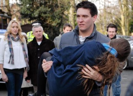 Man carrying an unconscious woman in his arms. Bystanders look on.