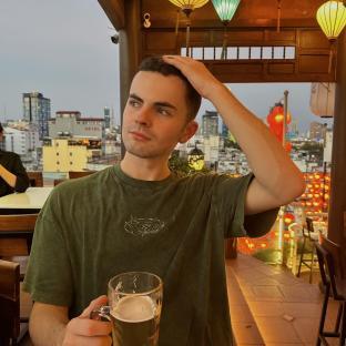 Man sitting outdoors at night, holding a beer, with a city skyline in the background.