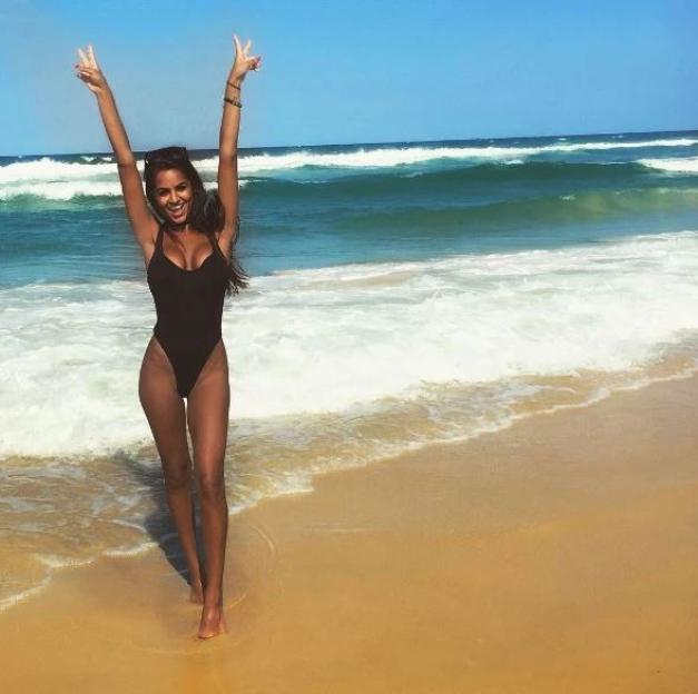 Danielle Scott posing in a black swimsuit on a beach with her arms raised, making peace signs with her hands.