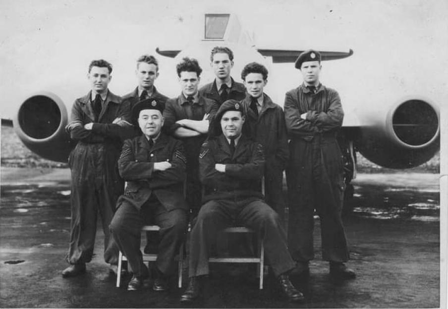 A group of RAF veterans, two seated and five standing, pose with their arms crossed in front of a military aircraft.