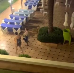 Video still of two women taking towels from sunbeds at a hotel pool at night.