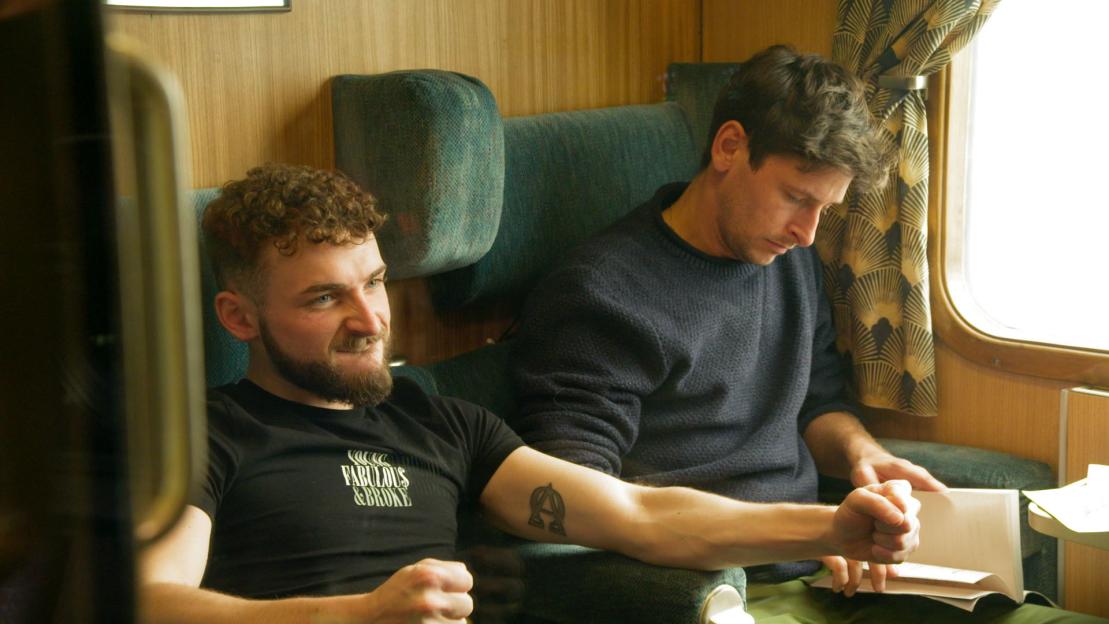 Two men sitting on a train, one reading a book.