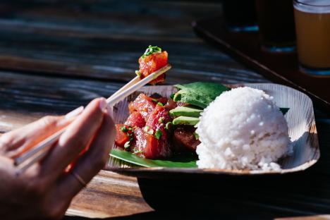 A person uses chopsticks to eat poke, served with rice and avocado.