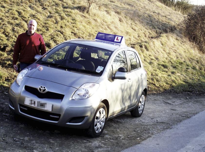 A man standing next to a silver Toyota Yaris with an L-plate on its roof.