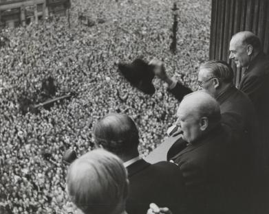 Black and white photo of Winston Churchill and other members of the cabinet addressing a large crowd from a balcony on VE Day.
