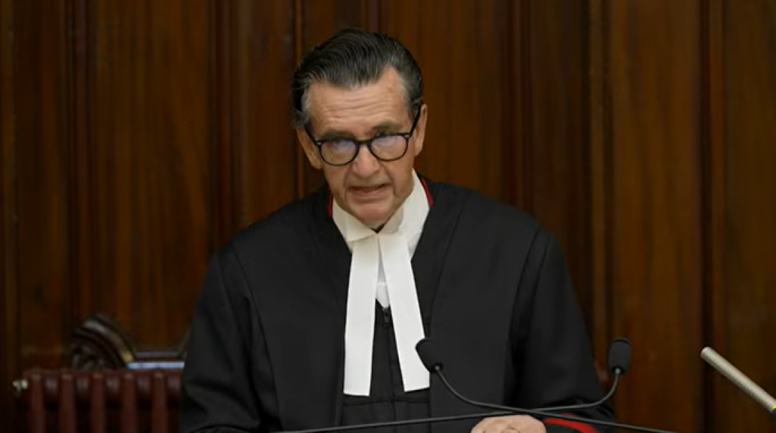 Judge in black robe and glasses speaking at a podium.
