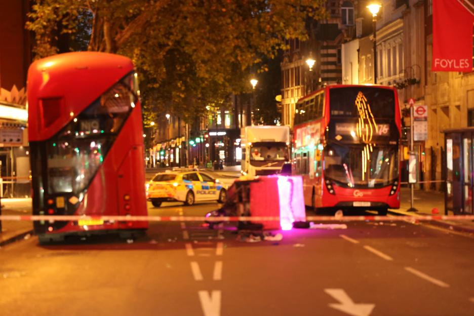 Nighttime photo of a damaged rickshaw at a London street accident scene, surrounded by police and emergency vehicles.