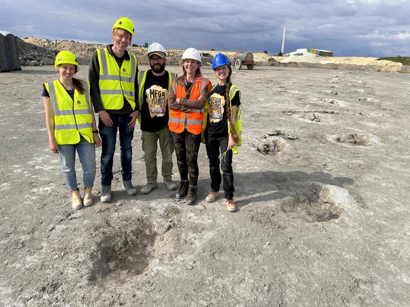 Five researchers in safety gear standing on an excavated site with multiple dinosaur footprints visible in the ground.