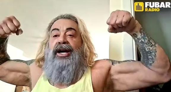 A muscular man with long gray hair and tattoos flexes his biceps for Fubar Radio.