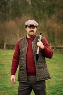 Man in tweed cap and hunting vest carrying a shotgun.