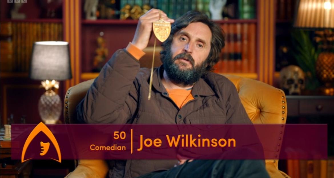 Joe Wilkinson displaying a golden pendant, with the text overlay "50 Comedian Joe Wilkinson".
