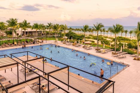 Hotel pool with many people swimming and relaxing on lounge chairs near the beach.