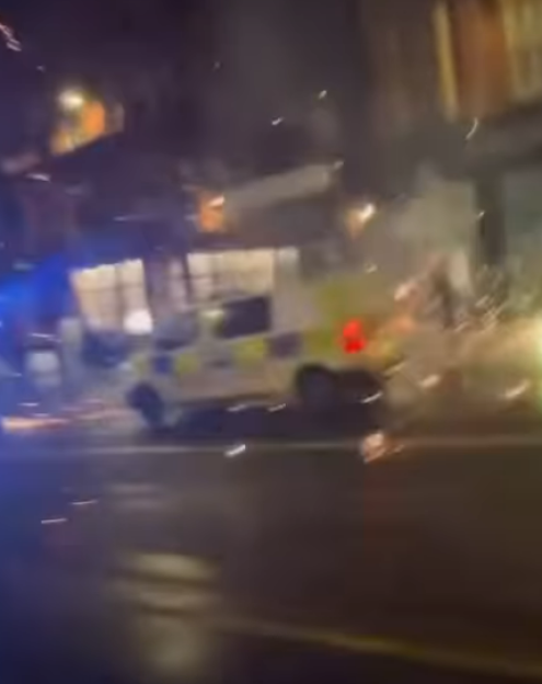 Police van with emergency lights on, surrounded by sparks, possibly from an explosion or fireworks.