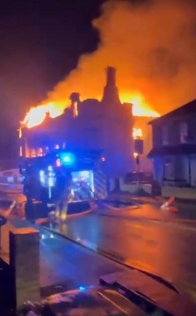 A large building engulfed in flames at night, with a fire truck in the foreground.