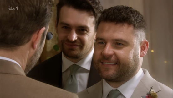 Screenshot of a groom smiling at his wedding, with two other men in suits in the background.