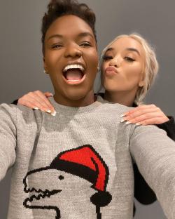 Two women posing for a selfie; one is wearing a grey sweater with a dinosaur wearing a Santa hat.