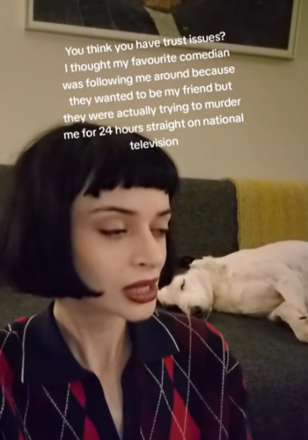 A woman with dark hair and red lipstick on a couch, with a dog sleeping behind her, and overlaid text that reads "You think you have trust issues? I thought my favorite comedian was following me around because they wanted to be my friend but they were actually trying to murder me for 24 hours straight on national television."