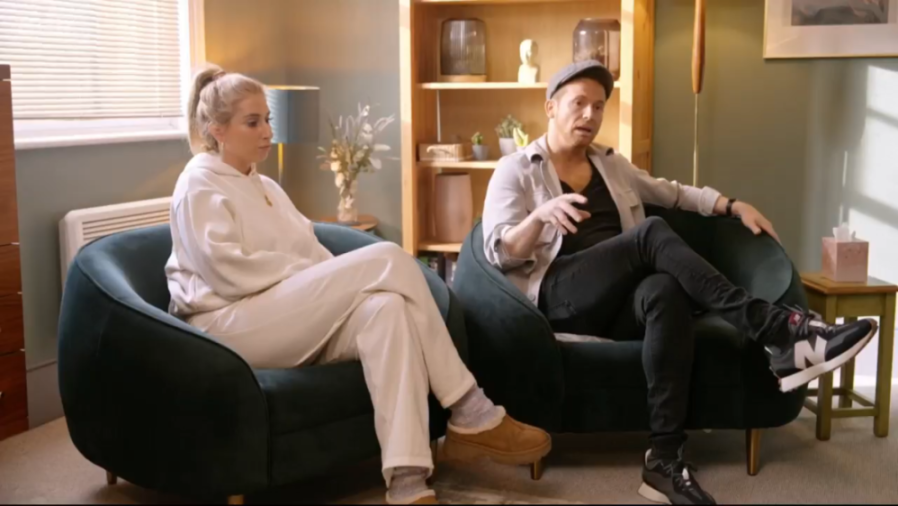 A man and a woman seated in dark blue armchairs, talking to each other.