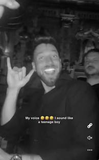 Black and white screenshot of a man making a hand gesture and smiling.