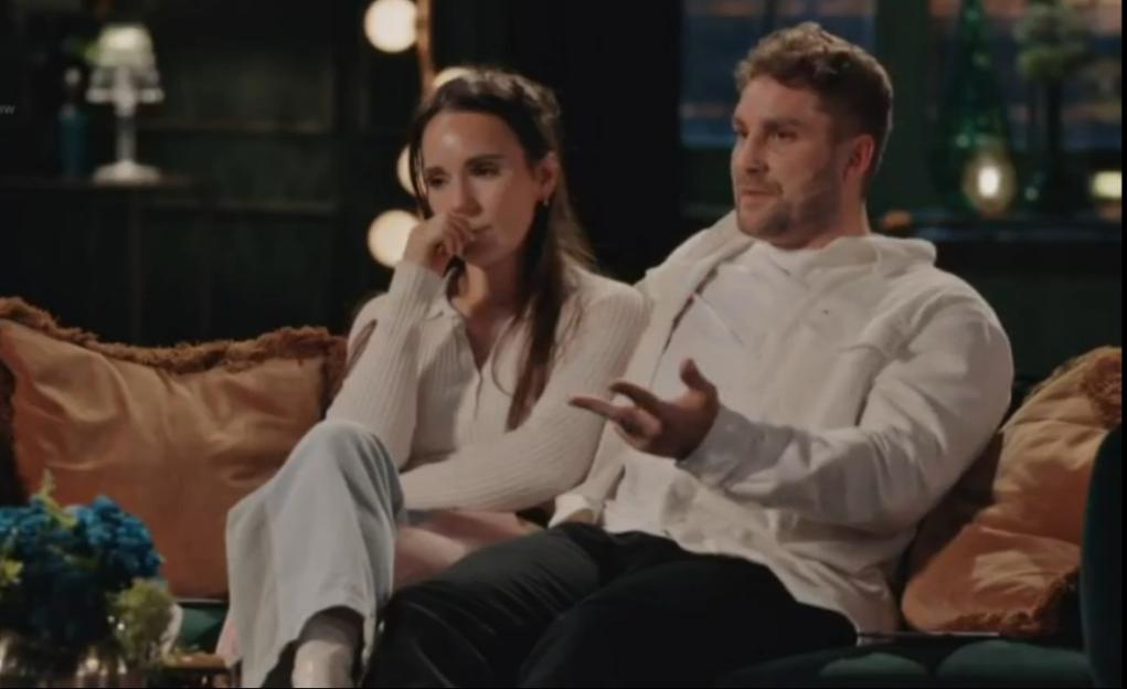A man and woman sitting on a dark green couch, the woman looking down and the man looking right, speaking.