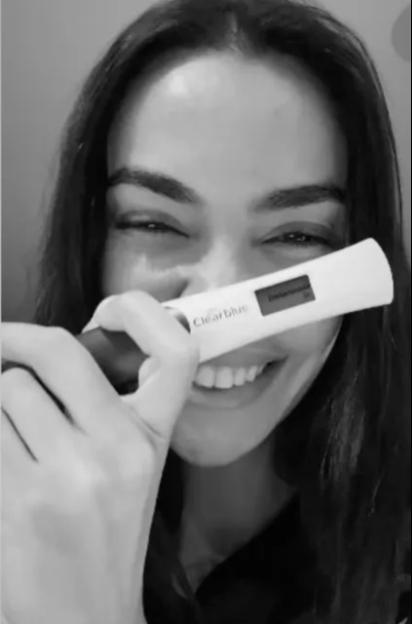 A woman holding a Clearblue pregnancy test that reads "Schwanger" and smiling widely.