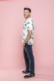 Man in floral shirt and jeans smiling against pink background.