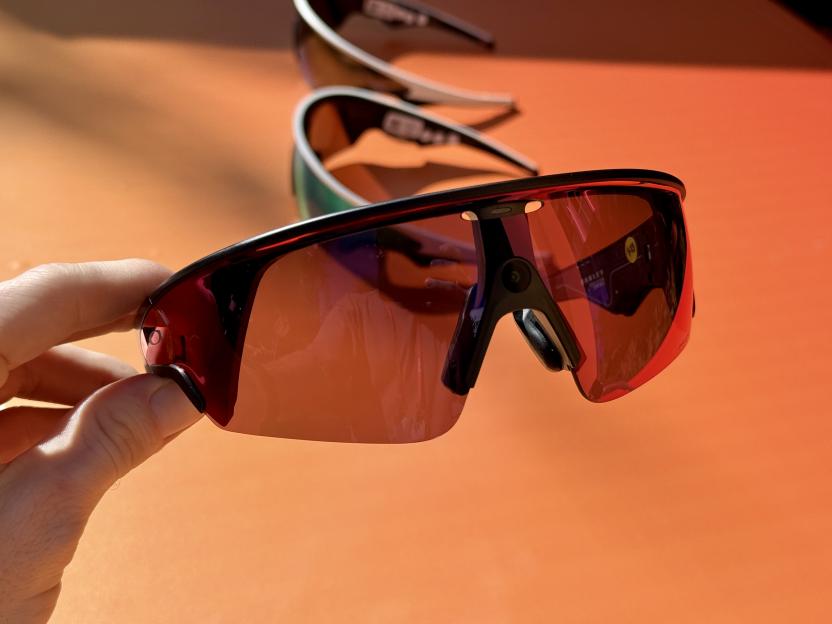 A hand holding a pair of sports sunglasses with orange lenses, with two other similar pairs blurred in the background on an orange surface.