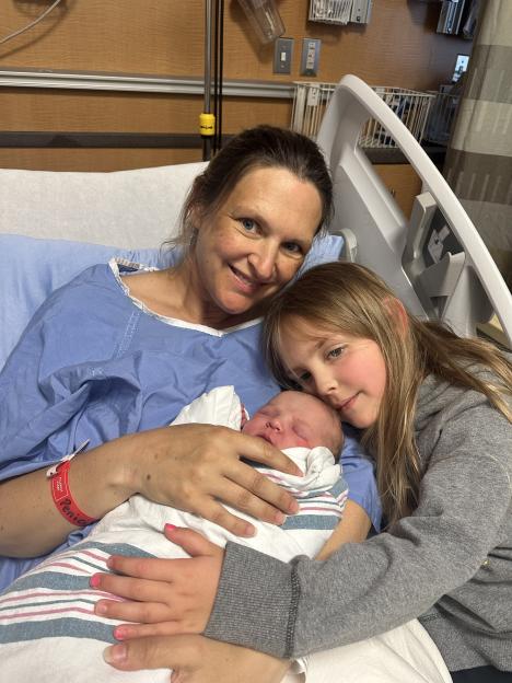 Woman in hospital bed holding newborn baby, with older daughter cuddling.