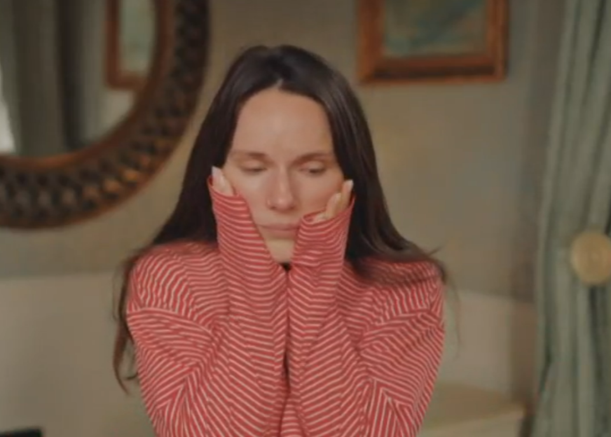 A woman in a red and white striped shirt with long sleeves pulled over her hands, holding her face, looks downcast.