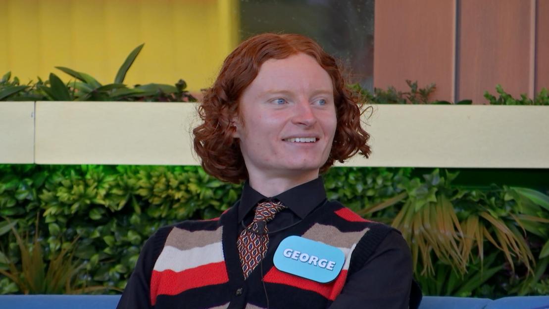 Contestant George in a black shirt, patterned tie, and striped cardigan smiling.