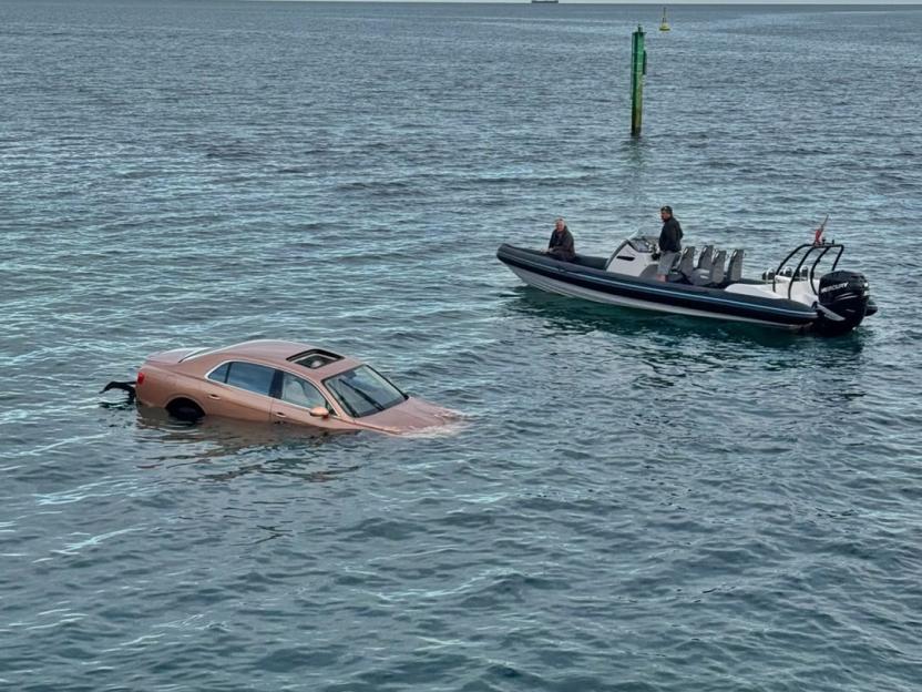 A bronze-colored car sinking in the sea next to a rescue boat.