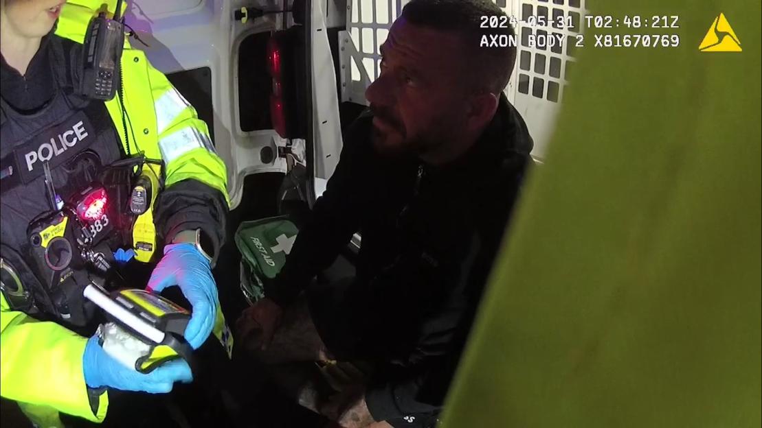 A police officer in a high-visibility jacket with the word "POLICE" on her vest, speaking to a man inside the back of a police van. The officer is holding a device while wearing blue gloves.