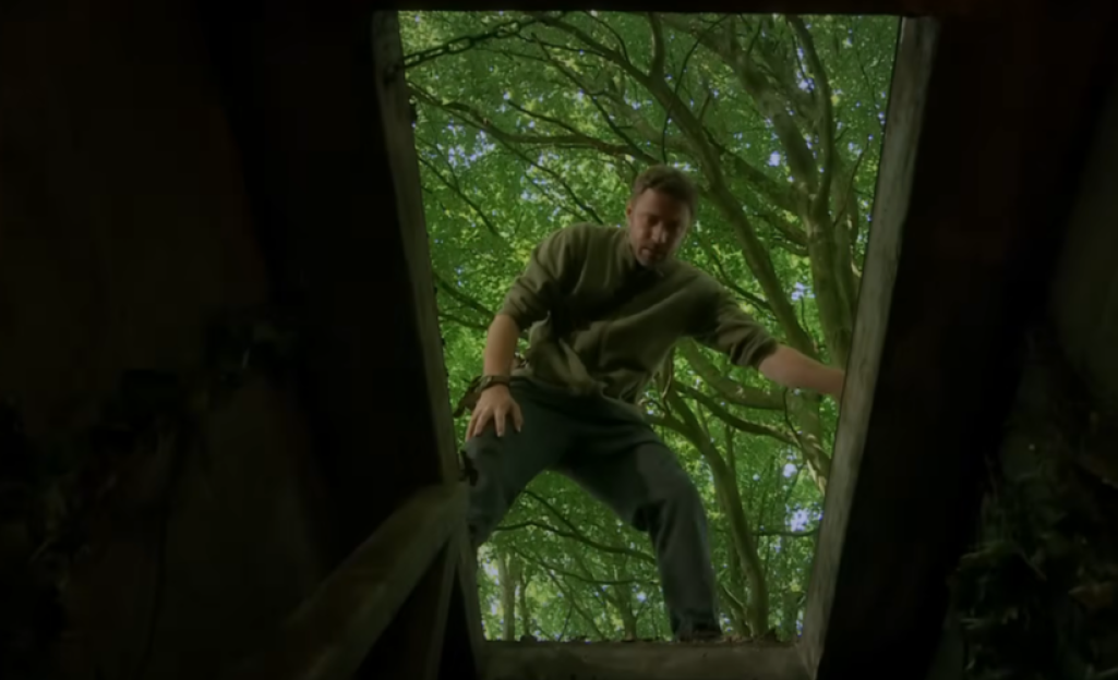 A man emerging from a shallow grave in a wooded area.