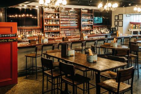 Interior view of Down One Bourbon Bar in Louisville, showing a long bar stocked with bourbon bottles and several wooden tables and chairs.
