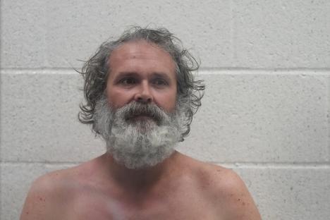 Shirtless man with long gray hair and beard against a white wall.