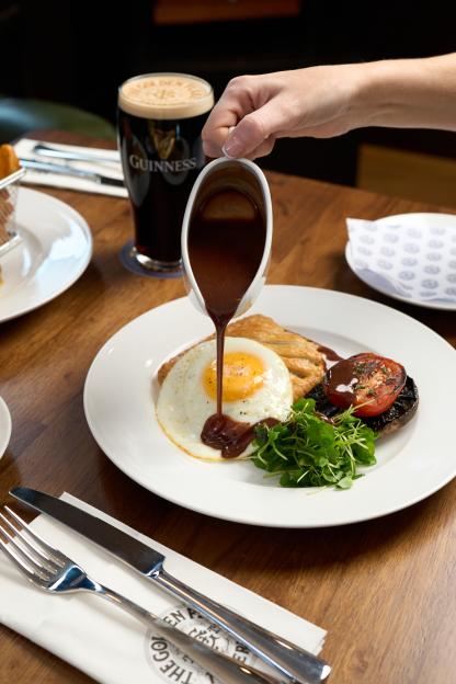 Hand pouring gravy over a full English breakfast with a Guinness pint in the background.