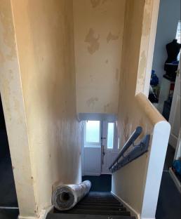 A rolled-up carpet sits on stairs leading to a door with damaged walls.