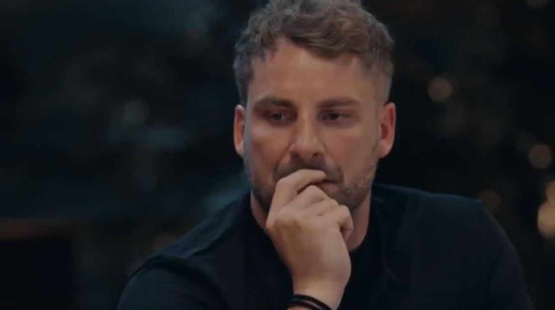 Man in a black t-shirt looking down, with his hand touching his chin.