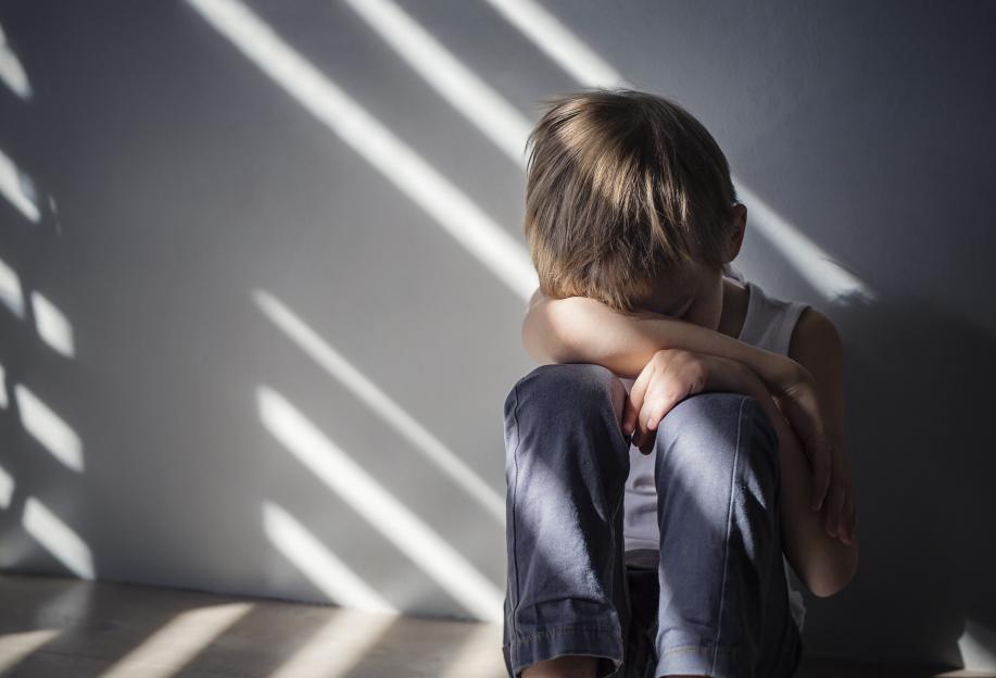 A sad, lonely, and depressed young boy sits with his head down, shadowed by window blinds.