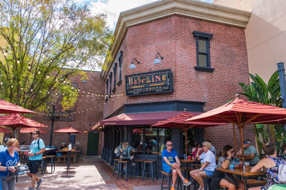 People seated at outdoor tables under red umbrellas outside The BaseLine Tap House bar at Walt Disney World Resort's Hollywood Studios.