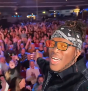 KSI wearing Simon Cowell's glasses in front of a large crowd.