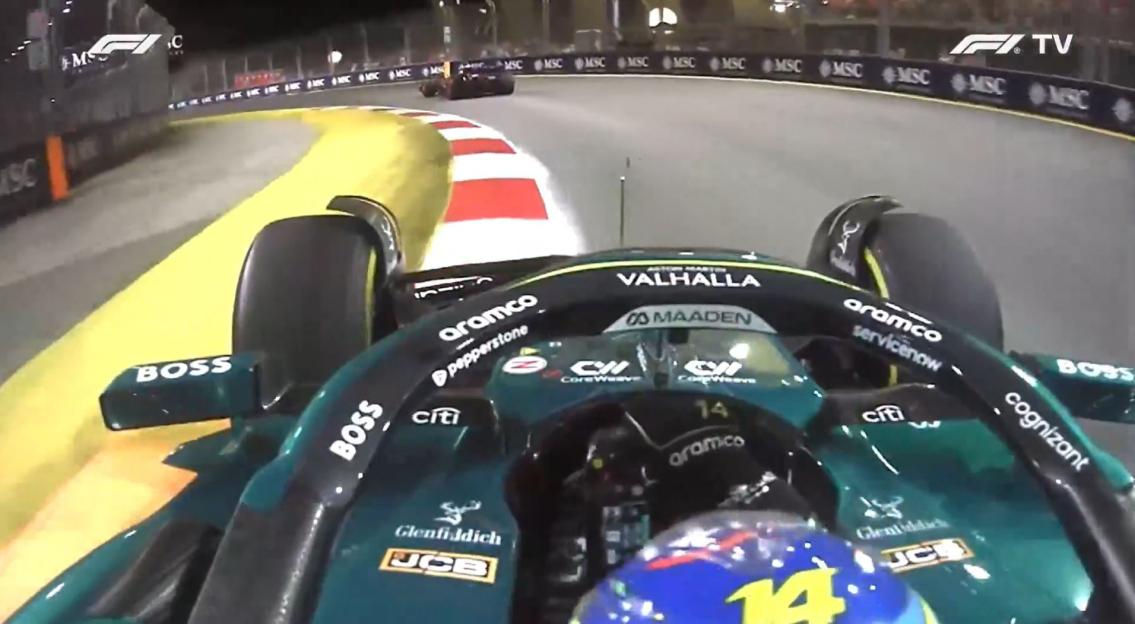 Onboard camera view from a dark green Formula 1 car during a night race, showing the front wing and the track ahead, with another car in the distance.