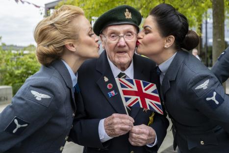 A 100-year-old World War II veteran is kissed on each cheek by two women in military-style uniforms as he holds a small Union Jack flag.