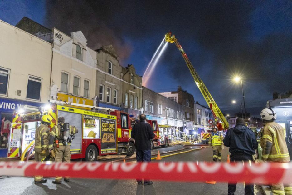 Firefighters battle a shop fire in Penge, London, with a ladder truck spraying water.