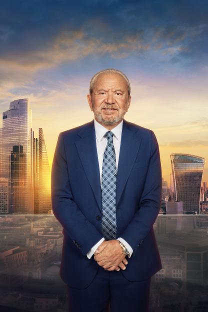 Lord Alan Sugar in a blue suit and tie, standing in front of a city skyline at sunset.