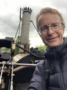 A man smiles for a selfie in front of a steam train.
